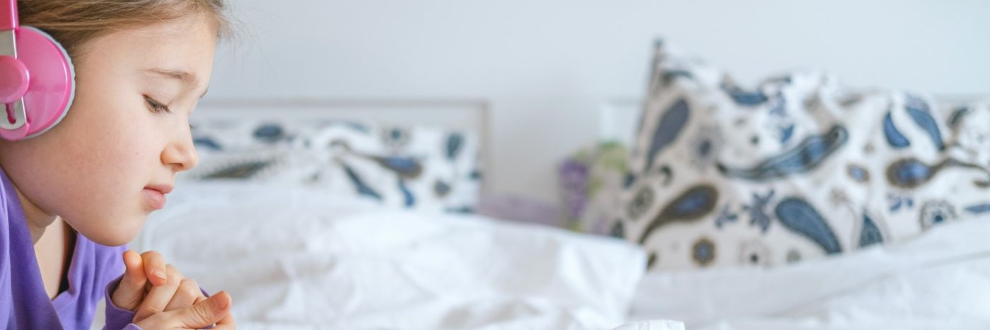 A girl with pink headphones listening to something, looking down, next to a bed with patterned pillows.