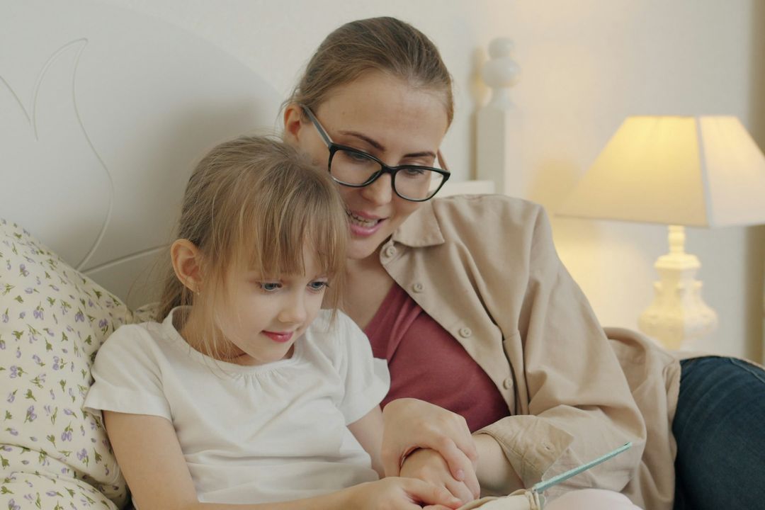 A mother and her daughter looking at a book together in bed, with a lamp on behind them.