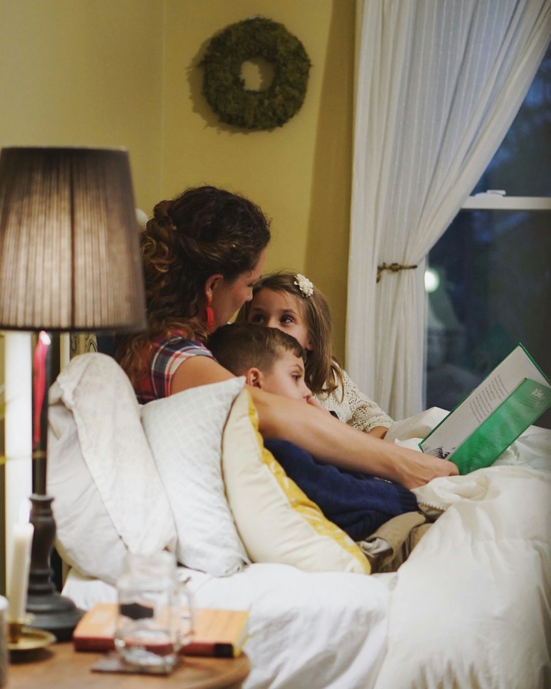 A mother reading to her two children in bed in the evening. The lamp is on and it is dark outside.